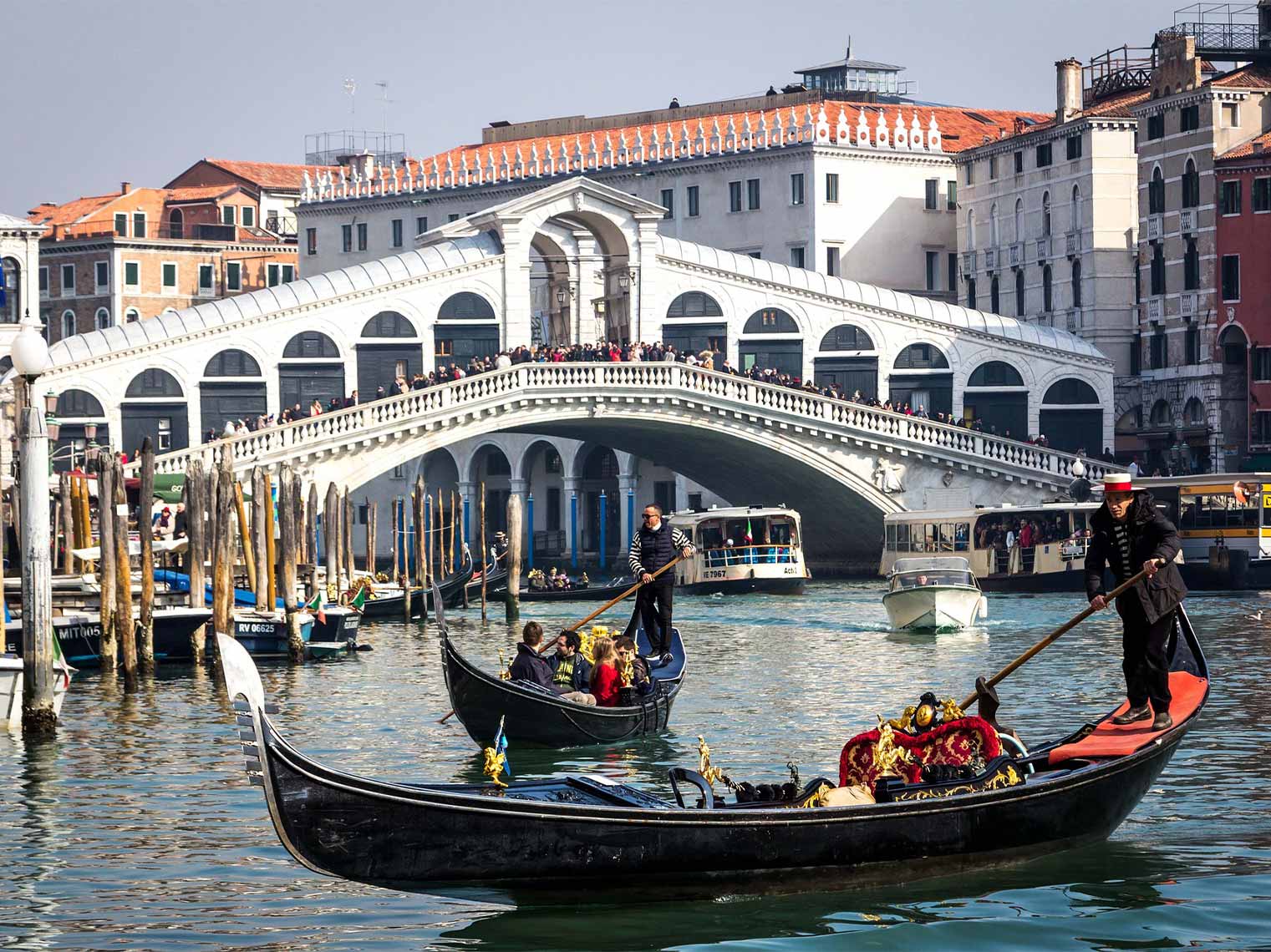 Guests enjoying a gondola tour in Venice while others walk over a white bridge.