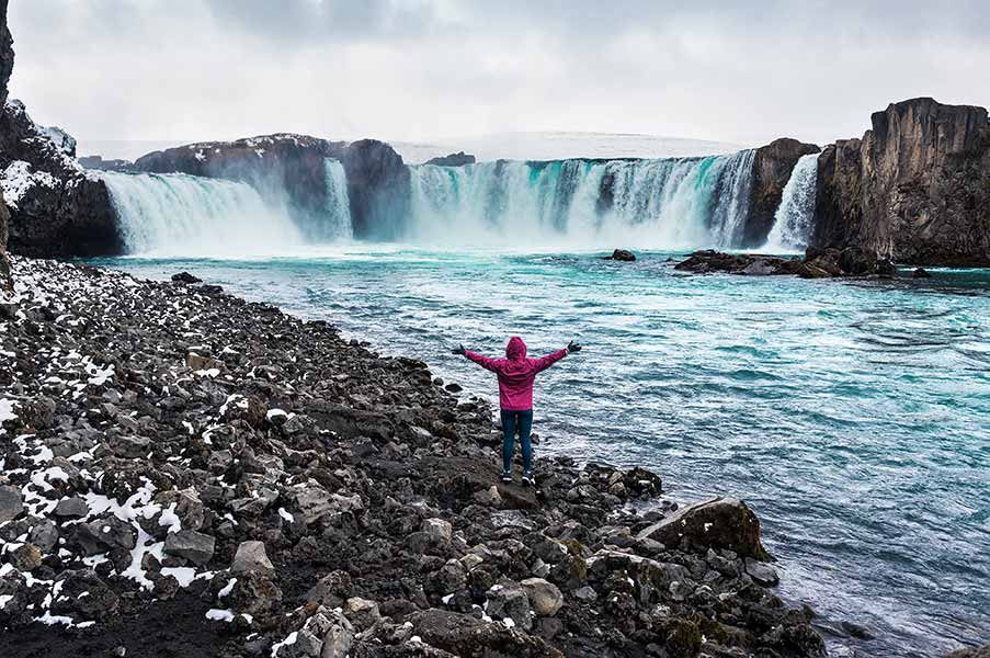 woman with arms outstretched in pink jacket at Godafoss Waterfall, Iceland