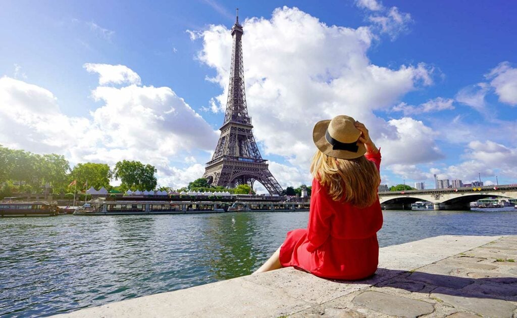Tourist in red dress sitting by the Seine River admiring the Eiffel Tower on a sunny day in Paris