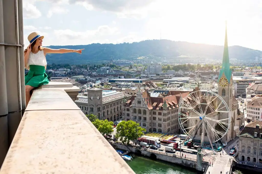 Tourist enjoying the panoramic view of Zurich cityscape, featuring landmarks like Fraumünster Church and a large Ferris wheel, from a high vantage point