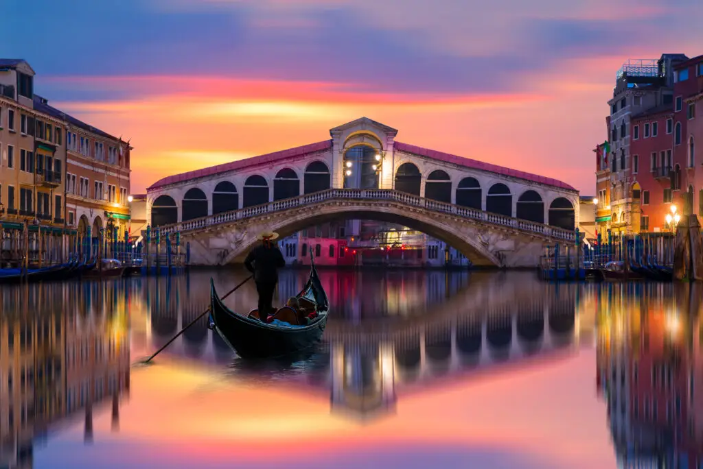 Gondola guided by a gondolier on the calm waters near the Rialto Bridge during sunset in Venice, Italy.