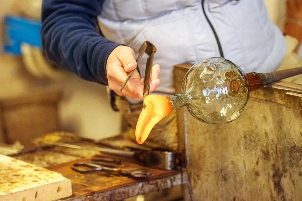 Master glassmaker in Venice, Italy, shaping a hot Murano glass blob using traditional tools, exemplifying skilled craftsmanship in Venetian glassmaking.