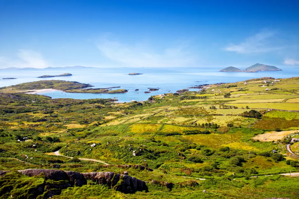 Panoramic view of the scenic Ring of Kerry in Ireland, featuring lush greenery, winding roads, serene Atlantic Ocean, and distant hills, a popular tourist destination.