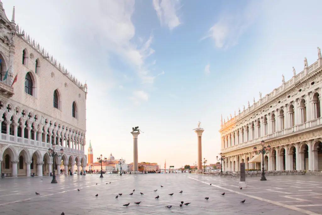 Early morning view of Doge's Palace and St. Mark's Column in Piazza San Marco, Venice, Italy, with clear skies and scattered pigeons on the square.