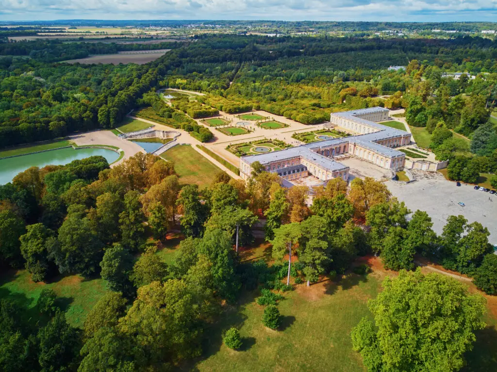 Aerial view of the Grand Trianon Palace surrounded by lush gardens in the Gardens of Versailles, Paris, France, showcasing the elaborate architecture and landscaped grounds.
