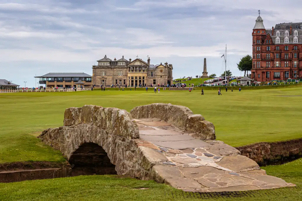 View of the historic Swilcan Bridge at St Andrews Golf Course in Scotland with golfers and the iconic clubhouse in the background