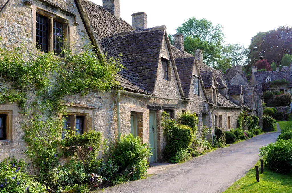 Charming view of traditional Cotswold stone houses with thatched roofs and lush gardens along a peaceful village road in the English countryside.