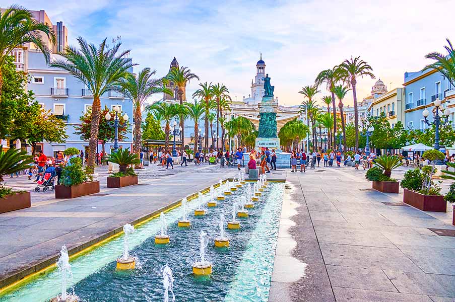 Vibrant view of Plaza de San Juan de Dios in Cadiz, Spain, featuring lively crowds, palm trees, and a historical monument with fountains under a clear blue sky.