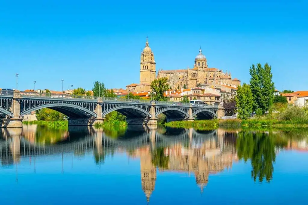 Panoramic view of the Salamanca Cathedral with Enrique Estevan Bridge over the tranquil River Tormes, reflecting the clear blue sky and historical architecture in Spain.