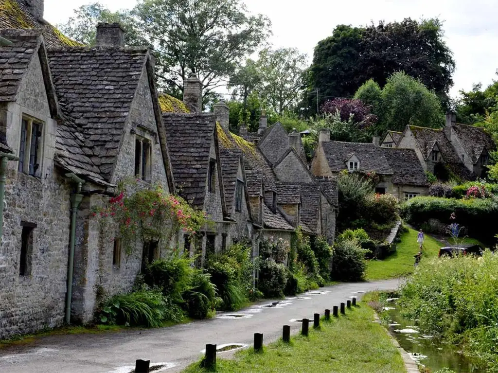 Scenic view of historic Cotswolds stone cottages with flowering gardens along a peaceful rural road in England, ideal for picturesque tours.