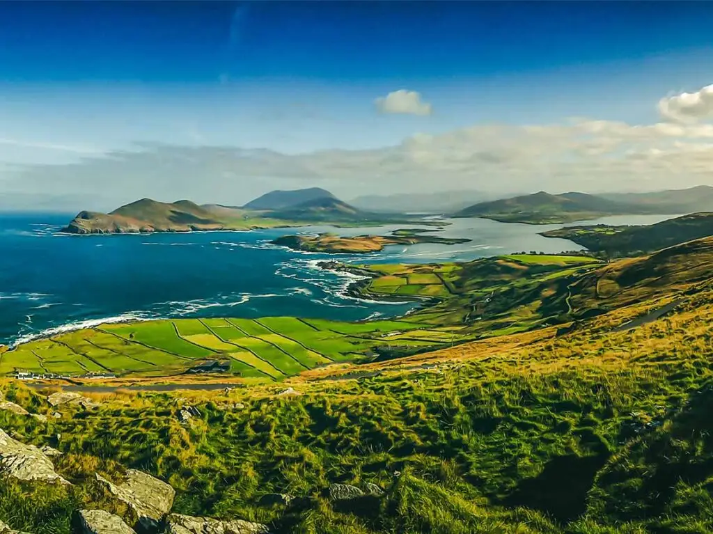 Panoramic view of County Kerry, Ireland showcasing rolling green hills, rugged coastline, and patchwork fields under a dramatic sky.