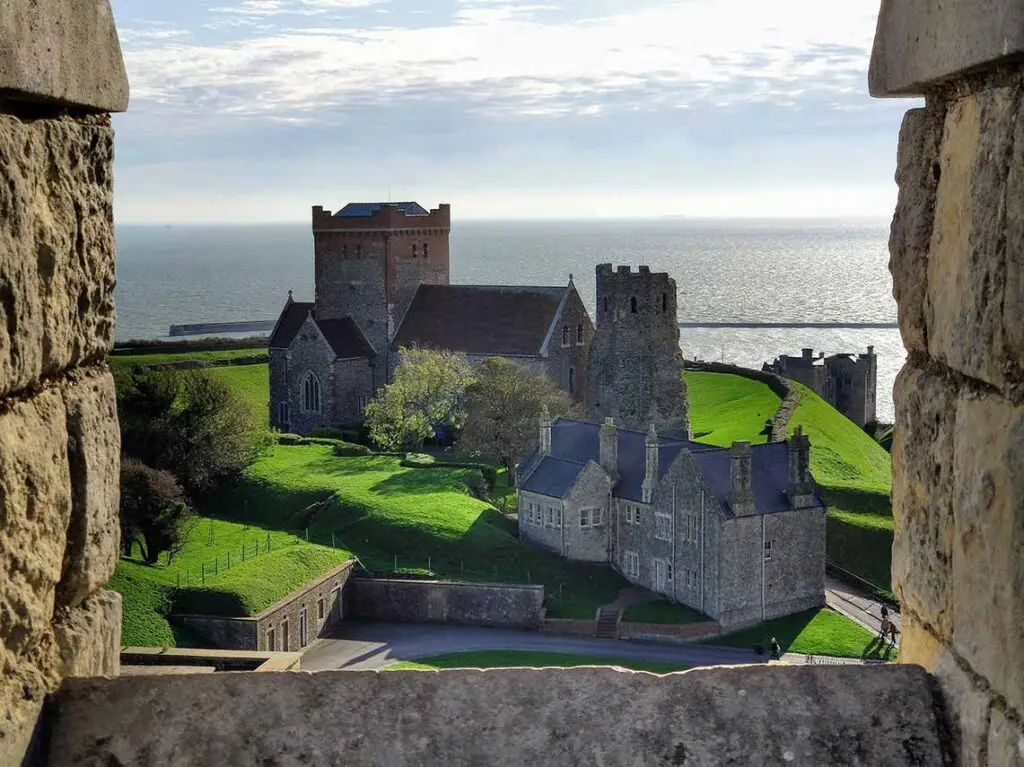View of Dover Castle and surrounding medieval buildings from a stone window, showcasing lush green lawns and the English Channel in the background, in Dover, England.