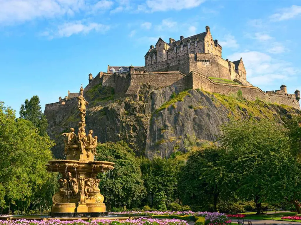 Majestic view of Edinburgh Castle perched on Castle Rock above Princes Street Gardens, showcasing the Ross Fountain in the foreground, under a clear blue sky in Scotland.