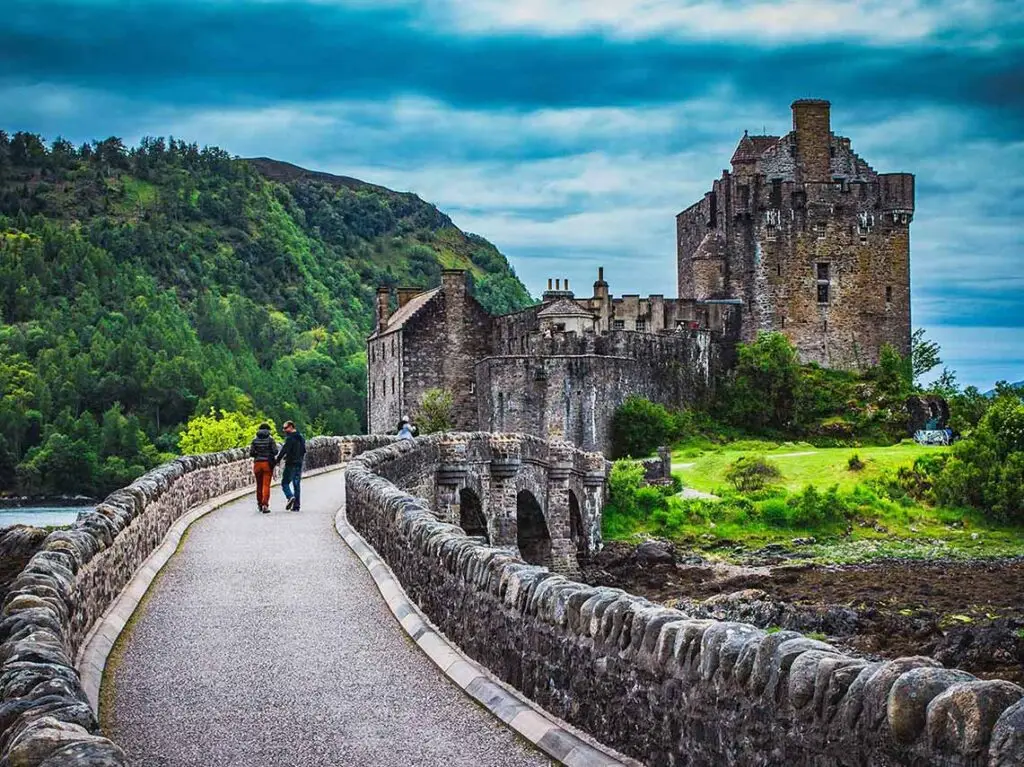 Visitors walking towards the historic Eilean Donan Castle in Scotland, showcasing lush greenery and dramatic stone architecture under a cloudy sky.