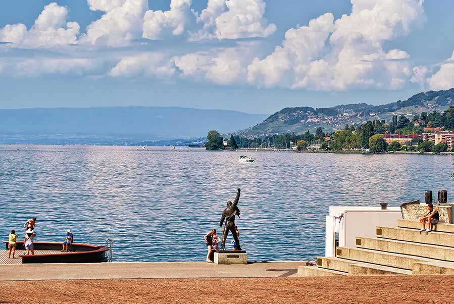 Freddie Mercury statue overlooking Geneva Lake in Montreux, Switzerland, with tourists enjoying the scenic lakeside view.