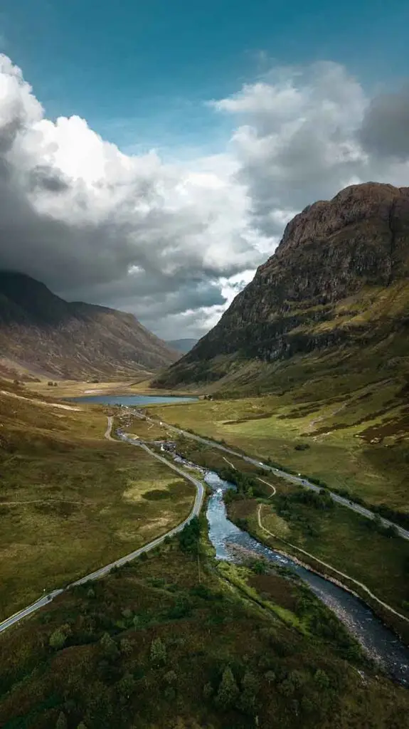 Breathtaking aerial view of Glencoe Valley in Scotland, showcasing lush greenery, winding river and roads under dynamic cloudy skies.