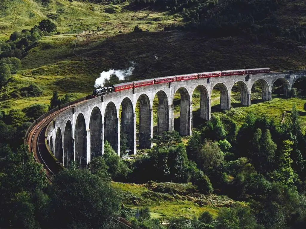 Vintage steam train crossing the iconic Glenfinnan Viaduct amidst lush green Scottish Highlands.