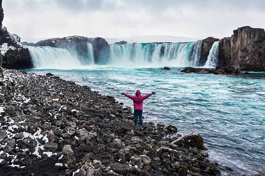 Tourist in pink jacket standing at Godafoss Waterfall in Iceland, admiring the majestic cascades and icy waters