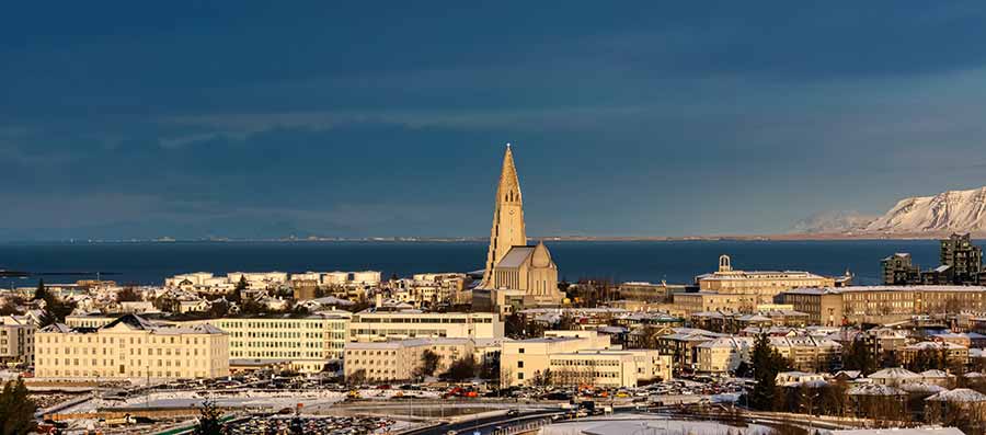 Panoramic view of Hallgrimskirkja church dominating the skyline of Reykjavik, Iceland, with snow-covered mountains in the background and cityscape in the foreground.
