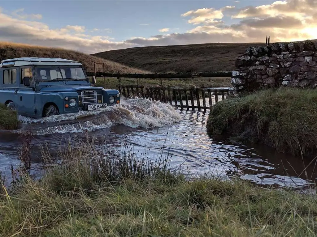 Vintage blue Land Rover crossing a flooded road in the Scottish Highlands during a safari tour, showcasing the rugged terrain and dramatic skies typical of Scotland.