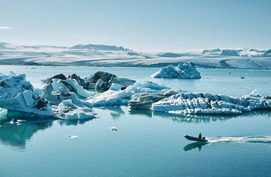 Tourist on a boat tour navigating the icy blue waters of Jokulsarlon Glacier Lagoon in Iceland, surrounded by floating icebergs and mountain views in the distance.
