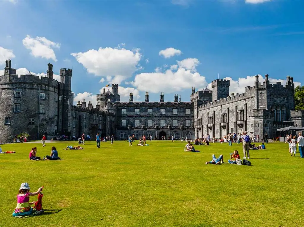 Visitors enjoying a sunny day on the lush green lawn in front of the historic Kilkenny Castle in Ireland.