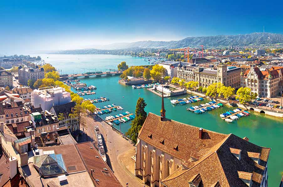 Panoramic view of Lake Zurich with vibrant cityscape and clear blue skies, showcasing boats docked in turquoise waters and historical buildings in Zurich, Switzerland.
