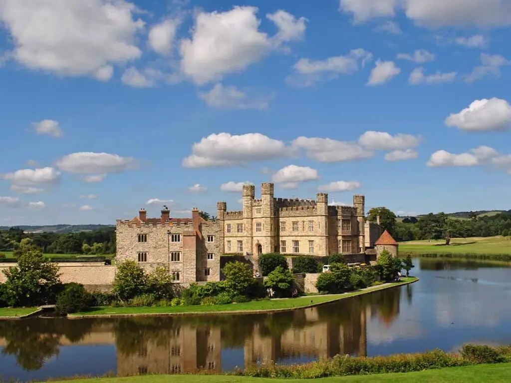 Scenic view of Leeds Castle in England with its historic stone architecture reflected in the water, surrounded by lush greenery and a clear blue sky.
