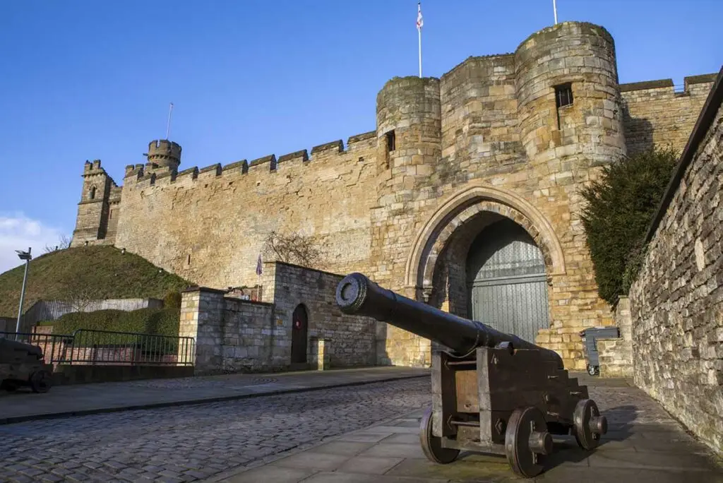 Historic Lincoln Castle in England with a foreground antique cannon, under a clear blue sky, showcasing medieval architecture and heritage tourism.