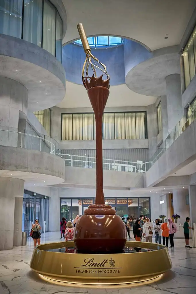 Giant chocolate fountain display inside Lindt Home of Chocolate in Zurich, Switzerland, with tourists admiring the exhibition in a modern architectural setting.