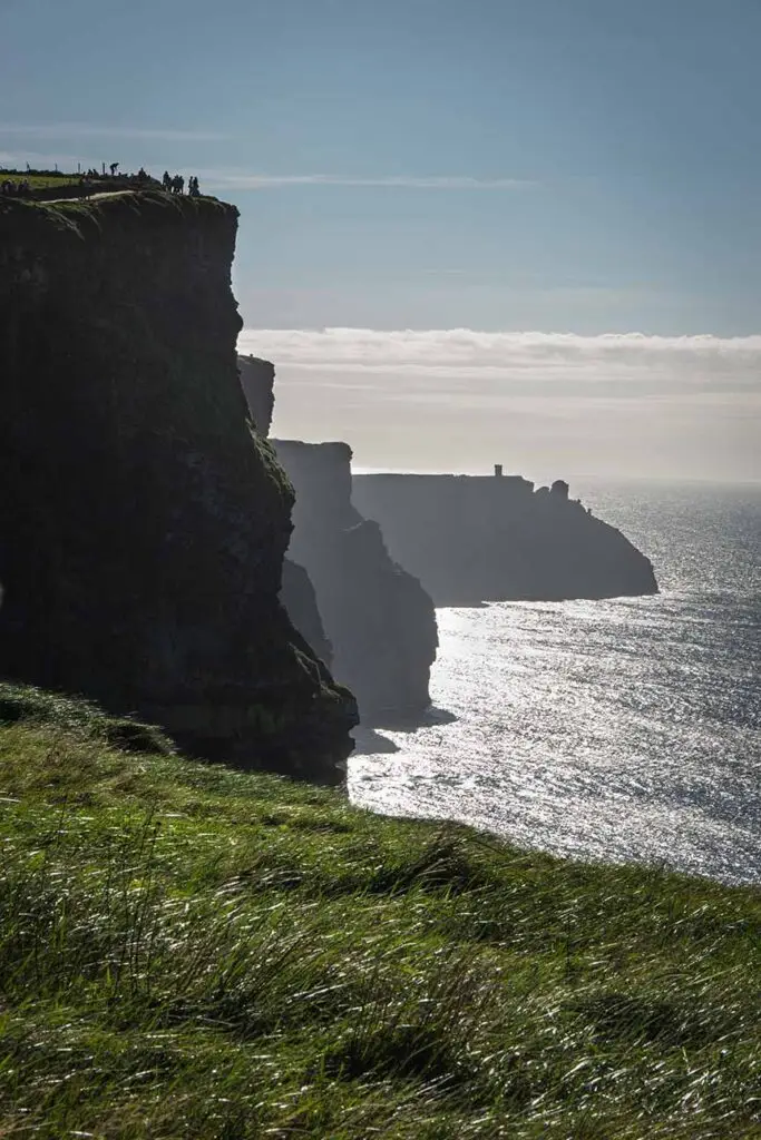 Misty view of the Cliffs of Moher in Ireland showing silhouetted tourists atop the grassy edge with the Atlantic Ocean gleaming under the sunlight.
