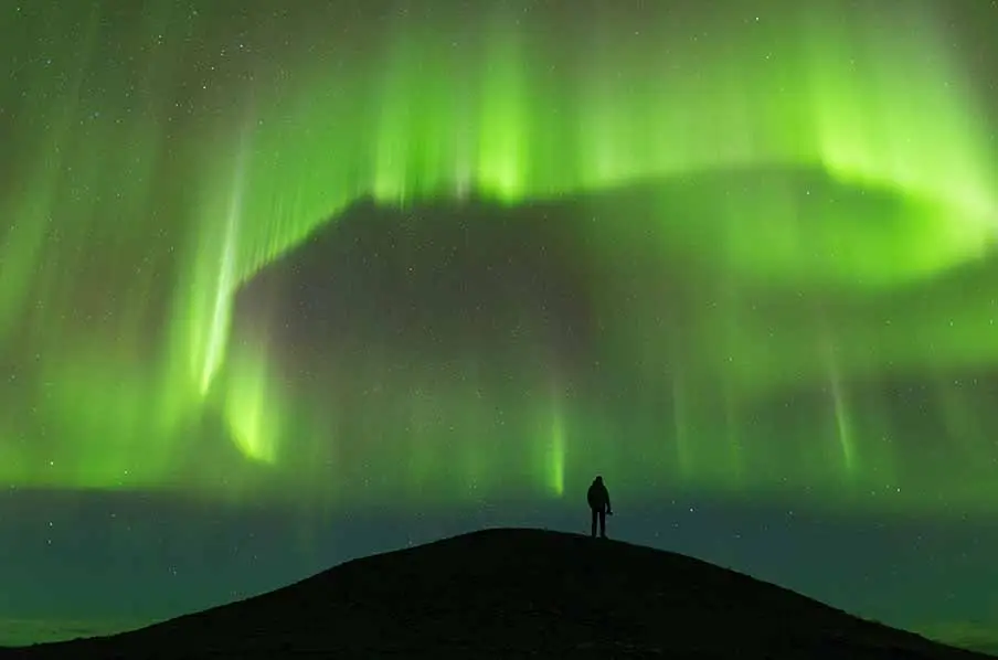 Silhouetted figure standing under the vibrant green auroras of the Northern Lights in Iceland.