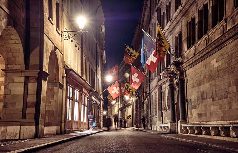 Nighttime view of a cobblestone street in Old Town Geneva, Switzerland, lined with historic buildings and Swiss flags.
