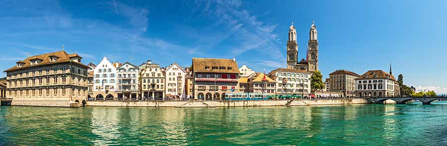 Panoramic view of Zurich's Old Town showcasing historical architecture along the Limmat River with the twin towers of Grossmünster Church prominent in the skyline.