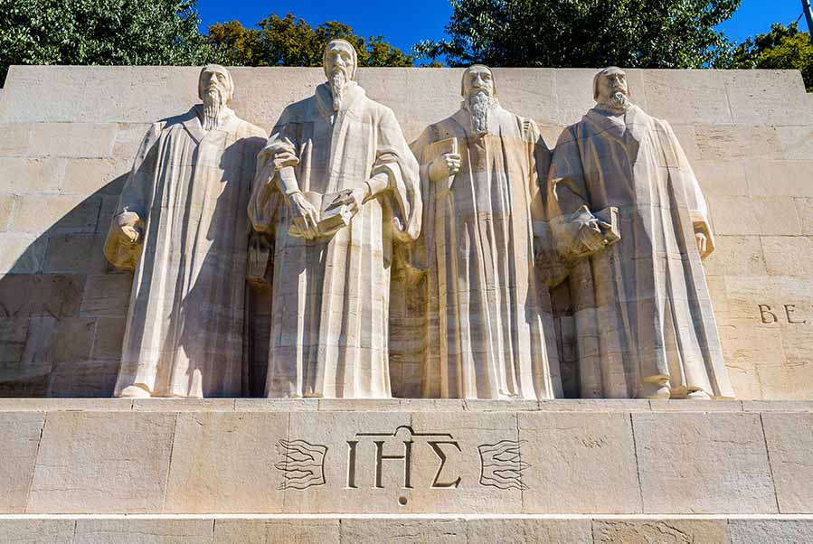 Sculptures of Protestant reformers at Reformation Wall in Geneva, Switzerland, depicting key figures in theological robes under blue sky.