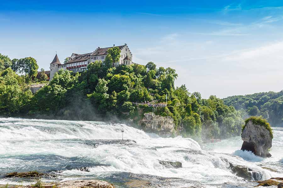 Majestic view of Rhine Falls in Switzerland, showcasing the powerful water cascades with a historic castle perched on lush green cliffs under a clear blue sky.