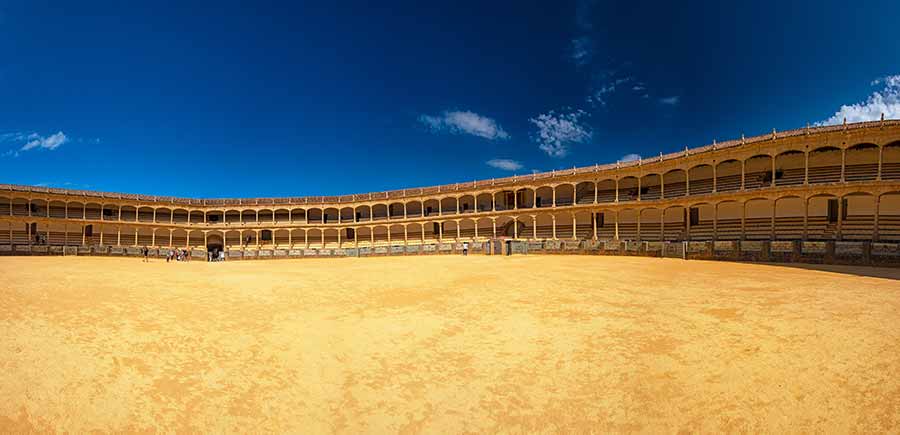 Panoramic view of the historic Ronda Bullring in Spain, showcasing its expansive sandy arena encircled by two-tiered seating under a clear blue sky.