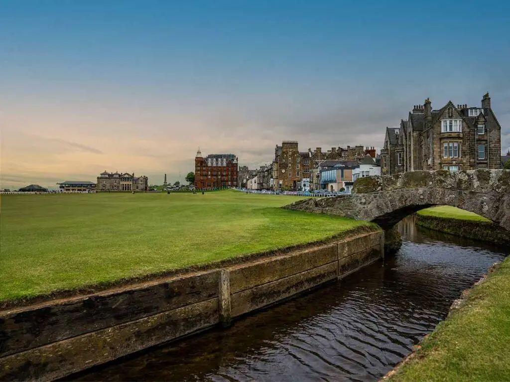 Scenic view of the iconic St. Andrews Old Course in Scotland with historic architecture and Swilcan Bridge over a serene waterway.