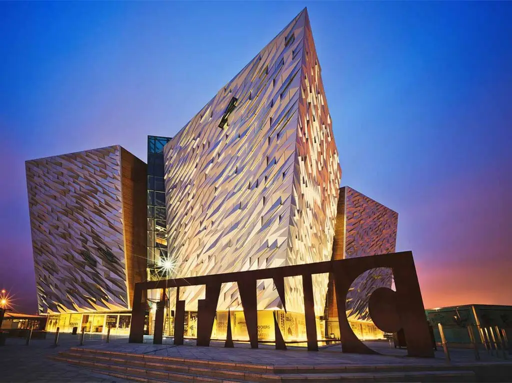 Titanic Museum Belfast at twilight with illuminated, geometrically designed building facade and prominent Titanic sign in the foreground.