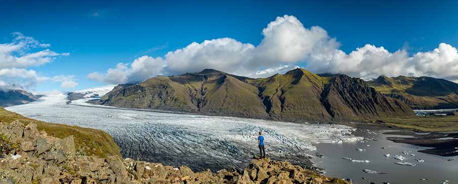 Panoramic view of a tourist observing the vast Vatnajokull Glacier in Iceland, showcasing dramatic mountain landscapes and expansive ice fields under a bright blue sky.